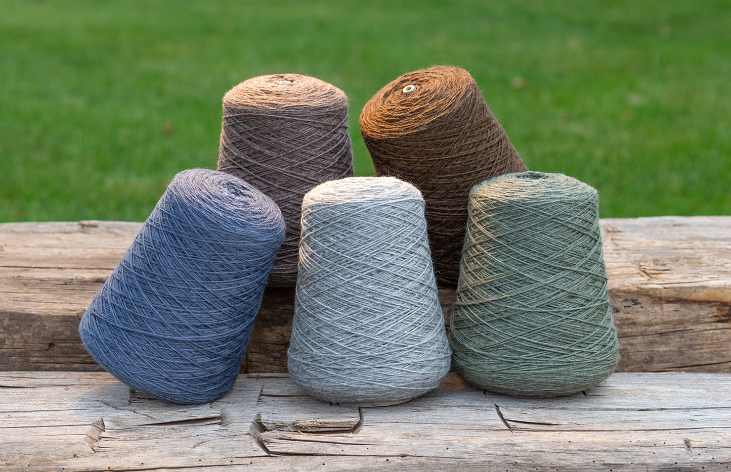 Four large cones of yarn in blue, gray, brown, and sage green, arranged on a rustic wooden surface with a blurred green grass background.